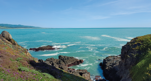 Imagem: Fotografia. Vista do alto de local com costa de rochas em cor cinza à esquerda e à direita, com grama de cor verde. Ao fundo, oceano de cor azul-claro com ondas de cor branca e rocha na água. Em segundo plano, à esquerda, morros. No alto, céu de cor azul-claro.  Fim da imagem.