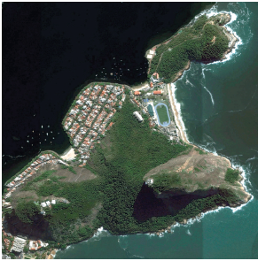 Imagem: Fotografia. Vista do alto de local com parte à esquerda em verde, pequena de moradias, ruas em cima. Na ponta da direita, partes rochosas em cinza e mais à direita, água do mar em verde.  Fim da imagem.