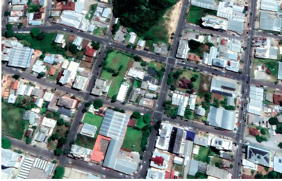 Imagem: Fotografia. Vista do alto de cidade com ruas de cor cinza-escuro, formando quadras compostas por construções, casas, gramado verde e partes com árvore de folhas em verde-escuro.  Fim da imagem.