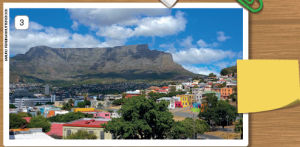 Imagem: Fotografia 3. Vista geral de local com cidade com casas de paredes coloridas e à direita, árvore de folhas verdes. Em segundo plano, morro alto de cor cinza e partes em tons mais escuros. No alto, céu em azul-claro com nuvens brancas.  Fim da imagem.