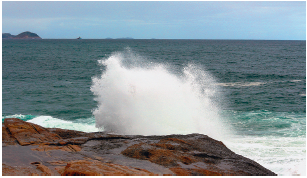 Imagem: Fotografia. À frente, rocha de cor marrom molhada e à frente, oceano de cor verde, batendo sobre rocha, jorrando água com gotículas branco. No alto, azul-claro sem nuvens.  Fim da imagem.
