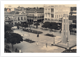 Imagem: Fotografia em tons de sépia. Vista do alto de praça, com árvores na ponta da esquerda e na ponta da direita. À direita, uma torre na vertical com um relógio redondo branco no alto. À esquerda, prédios antigos grandes, com telhado escuro. Fim da imagem.