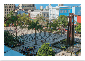 Imagem: Fotografia. Vista do alto da mesma praça descrita anteriormente, com árvores à esquerda e à direita, com muitas pessoas perto da árvore da esquerda sentadas e à direita, uma torre na vertical de cor marrom com um relógio redondo pequeno na parte superior. À esquerda, ao fundo, prédios de cor bege e casarões em tons de cinza claro e outros em azul. Em segundo plano, prédios modernos vistos parcialmente. No alto, céu azul-claro.  Fim da imagem.