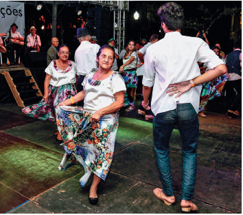 Imagem: Fotografia. Duas pessoas dançando, um homem visto de costas de camisa de cor branca, até os ombros, com calça jeans azul, fazendo passo de dança com a mão direita para trás. À esquerda, uma senhora mulher de pele clara, cabelos castanhos, com blusa de mangas curtas em branco e saia de estampas com flores e pétalas laranjas e folhas verdes, fazendo passo com as mãos na saia.  Fim da imagem.