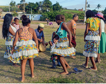 Imagem: Fotografia. Vista de local aberto com grama seca, algumas em verde, com mulheres e homens dançando. À frente, duas mulheres com saias curtas e à direita, com saia longa estampada. A mulher da esquerda, com blusa de laranja sem mangas, outra mulher sem mangas em azul e outra com mangas curtas em amarelo e estampa, vistas de costas, dançando. Mais ao fundo, uma mulher em pé de pele morena, cabelos pretos longos, com o mesmo tipo de roupa das outras, segurando uma criança no colo, desfocada. Ao lado, um homem agachado tocando tambor, de pele morena, blusa azul, com chapéu bege na cabeça; e outros homens. Em segundo plano, vegetação rasteira verde e árvores de folhas verdes, moradias e postes de energia. No alto, céu azul-claro com nuvens.  Fim da imagem.