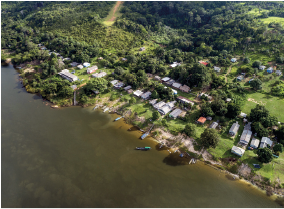 Imagem: Fotografia. Visto alto de local com rio de água escura à frente e na borda, embarcações pequenas perto do rio. À frente, local com grama e árvores de folhas verdes, com casas entre elas. Fim da imagem.