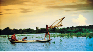 Imagem: Fotografia. Vista geral de local com rio escuro e sobre ele, canoa fina na horizontal onde há três pessoas. Na ponta da esquerda, um homem moreno de cabelos escuros, sem camiseta, sentado. Ao centro, um menino moreno de cabelos escuros com o corpo curvado para frente e na ponta da direita, um homem em pé, sem camiseta e short preto, jogando com as mãos uma rede para frente. Ao redor do rio, vegetação verde flutuando. Em segundo plano, árvores de folhas em verde-escuro e no alto, céu azul-claro com tons em marrom-claro e à direita, nuvem branca.  Fim da imagem.