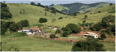 Imagem: Fotografia. Vista geral de local com vegetação em verde-claro, árvores com folhas em verde-escuro e ao centro, uma casa de paredes de cor branca, com telhado em bege-claro. Em segundo plano, morros com vegetação verde e árvores de folhas em verde-escuro. No alto, céu em azul-claro.  Fim da imagem.