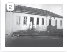 Imagem: Fotografia 2 em preto e branco. Uma casa de paredes de cor clara, telhado no alto de cor escuro. Nela há janelas pequenas e ao centro, de frente para uma porta, em cima do primeiro degrau de uma escada: duas pessoas. À esquerda, uma mulher de vestido claro com mangas curtas e à direita, sobre cadeira, uma pessoa sentada.  Fim da imagem.