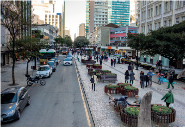 Imagem: Fotografia. Vista geral de local com uma rua à esquerda, de cor cinza, com carros estacionados à esquerda e à direita, outros carros passando. Mais à direita, calçada por onde pessoas passam, com bancos de madeira e vegetação com cerda em madeira. Nas extremidades, prédios de paredes claras e em segundo plano, prédios altos de cor cinza.  Fim da imagem.