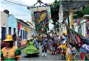 Imagem: Fotografia. Ao centro, rua de cor cinza e nas pontas extremas, casas de paredes coloridas uma ao lado da outra. Na rua, dezenas de pessoas passando, fantasiadas com roupas coloridas. À esquerda, mulher de vestido longo verde e ao lado dela, um homem de roupa de mangas compridas em azul-escuro com estampas dourada e mais atrás, no alto, um tecido pendurado no alto com desenho de um peixe dourado, com o título: Piaba de ouro 2018 e um homem e tons de dourada, com camiseta vermelha e bermuda azul, tocando tambor. No alto, fios de energia preta, com céu em azul-claro.  Fim da imagem.