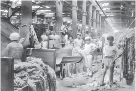 Imagem: Fotografia em preto e branco. Dentro de uma fábrica, com hastes finas na vertical, vê-se pessoas, em meio a máquinas, tecidos e linhas. À esquerda, local com linhas emaranhadas. Mais ao fundo, ao centro, homem em pé, olhando para frente, com boina sobre a cabeça, camiseta clara e calça, com a mão direita sobre a cintura. Mais ao fundo, à esquerda, outros homens em pé e estruturas na vertical, com base na horizontal com tecidos.    Fim da imagem.