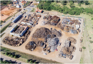 Imagem: Fotografia. Vista do alto de local com terreno grande, com solo em marrom-claro, montes de terra de cor marrom, com ferros de cor cinza, marrom espalhados entre os solos. Ao redor, local arborizado de folhas de cor verde.  Fim da imagem.