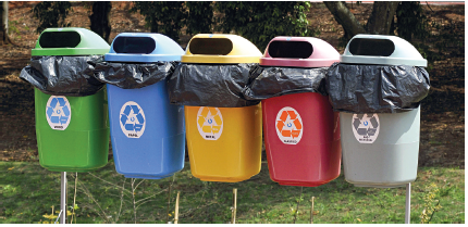 Imagem: Fotografia. Penduradas no alto, cinco lixeiras coloridas com sacola preta dentro, com símbolo de reciclagem. Da esquerda para à direita: em verde, em azul-claro, em amarelo, em vermelho e em cinza. Ao fundo, grama verde e árvores de troncos marrons e vegetação.  Fim da imagem.