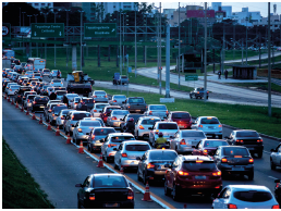 Imagem: Fotografia. Vista do alto de local com avenida extensa de cor cinza por onde passam dezenas de carros no tráfico, um atrás do outro.  Fim da imagem.