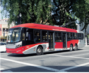 Imagem: Fotografia. Sobre uma rua de cor cinza, com linhas brancas, com ônibus de cor vermelha, janelas e na parte inferior, partes em cinza-claro e detalhes em branco. Na parte superior, árvore de folhas verdes, céu de cor azul-claro.  Fim da imagem.