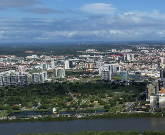 Imagem: Fotografia. Vista do alto de local com rio na horizontal de rio de água escura, com bordas de cor verde. Mais ao fundo, local com vegetação e árvores de folhas verdes. Em segundo plano, cidade com muitos prédios de tamanhos médio e altos, a maioria de paredes de cor cinza-claro. No alto, céu em azul-claro com muitas nuvens brancas.  Fim da imagem.