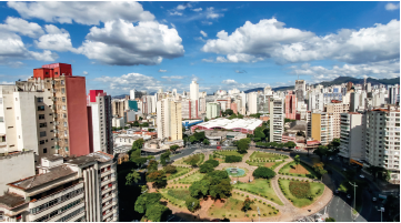 Imagem: Fotografia. Vista do alto de cidade composta por prédios à esquerda e à direita, de tamanhos médios e alguns mais altos. A maioria tem paredes de cor branca, cinza-claro e um prédio à esquerda de paredes de cor avermelhada. Ao centro, local com ruas de cor cinza, com vegetação rasteira e algumas árvores de folhas verdes. Mais ao fundo, um grande galpão baixo e telhado de cor branco. No alto, céu de cor azul-claro com nuvens brancas.  Fim da imagem.