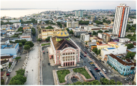 Imagem: Fotografia. Vista do alto do teatro descrito anteriormente. Ao centro, o teatro de telhado marrom, cúpula laranja e na parte inferior, paredes de cor branca com partes em bege. À frente, local com grama verde. À esquerda e à direita, prédios, casas e estabelecimentos.  Fim da imagem.