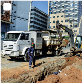 Imagem: Fotografia. À esquerda, calça cinza, caminhão de cor branca, parte inferior em aberto e perto, um guindaste de cor cinza de trator visto parcialmente. Na ponta da rua, um homem em pé com uniforme de cor azul-escuro de mangas compridas e calça. À direita, rua com buraco e solo de cor marrom, com vista parcial de outra pessoa com roupa azul. Fim da imagem.