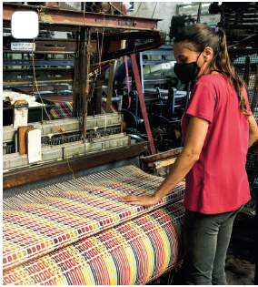 Imagem: Fotografia. À esquerda, máquina de tecelagem com hastes de cor marrom e à frente, tecido colorido em amarelo, partes em vermelho e roxo. Perto do tecido, uma mulher em pé vista de costas, com pele morena, com cabelos castanhos amarrados para trás, máscara no rosto de cor preta, com camiseta de cor vermelha-escura, com calça de cor cinza-escuro, com a mão esquerda sobre tecido. Fim da imagem.