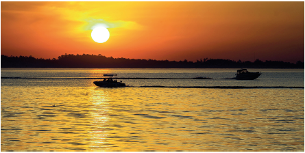 Imagem: Fotografia. Vista geral de local com mar à frente, de água escura, um barco pequeno à esquerda e outro mais ao fundo, à direita. Em segundo plano, céu de cor laranja com som redondo amarelo.  Fim da imagem.