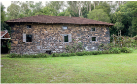 Imagem: Fotografia. À frente, local com grama verde, uma casa de paredes de pedra cinza e tons de bege, com pequenas janelas brancas e acima, telhado de cor marrom-escuro. Ao fundo, árvores de tronco fino e folhas verdes em cima.  Fim da imagem.