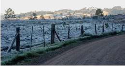 Imagem: Fotografia. À esquerda, local cercado de madeira, com vegetação rasteira coberta por neve branca, árvores ao fundo. À direita, local com solo marrom. Em segundo plano, morros e céu de cor cinza-claro.  Fim da imagem.