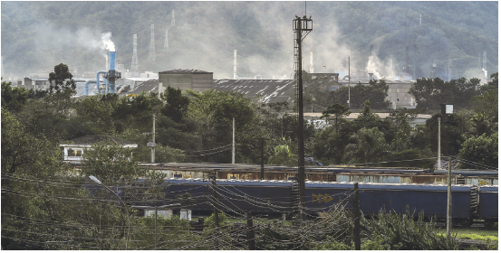 Imagem: Fotografia. Vista geral de local de vegetação de folhas verdes, vistas do alto. Ao centro, uma torre em ferro na vertical. Em segundo plano, galpões, tetos em cinza-escuro e entre eles, torres por onde sai fumaça branca em direção ao céu cinzento.  Fim da imagem.