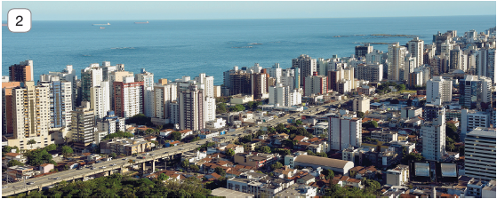 Imagem: Fotografia 2. Vista geral de cidade composta por prédios de tamanhos diferentes, baixo, médio e altos. Ao centro, casas e construções baixas, com rodovia sobre ponta à esquerda por onde passam carros. Em segundo plano, oceano de cor azul e no alto, céu em azul-claro.  Fim da imagem.