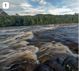 Imagem: Fotografia 1. À frente, local com pedras de cor cinza-escuro, com água de cor marrom, com ondas em tons de branco e marrom-claro. Em segundo, árvores de folhas verdes e morros. No alto, céu em azul com nuvens brancas. Fim da imagem.