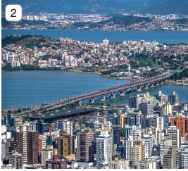 Imagem: Fotografia 2. Vista geral de cidade de prédios de tamanhos diferentes em branco, alguns em bege e outros em marrom. Ao fundo, ponte gigante de cor cinza com estruturas na vertical na base. Em segundo plano, local com vegetação em verde, prédios da cidade. Mais ao fundo, mar de água azul e no fundo, outra cidade com morros com partes em verde e prédios.  Fim da imagem.