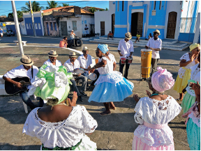 Imagem: Fotografia. Vista do alto de rua de cor cinza com dançarinas à direita, vistas de costas, e à esquerda, músicos sentados tocando instrumentos. As mulheres são de pele negra, vestido branco e na cabeça, lenço colorido em verde, azul, rosa-claro. Ao centro, uma delas está dançando, fazendo passo com os braços abertos. Os músicos são de pele negra, usam camiseta de cor branca, chapéu bege sobre a cabeça. Em segundo plano, rua cinza e em outra calçada, casas antigas de parede de cor azul, branco e à direita, em azul-claro. No alto, céu em azul-claro, com vista parcial de árvores com folhas verdes.  Fim da imagem.