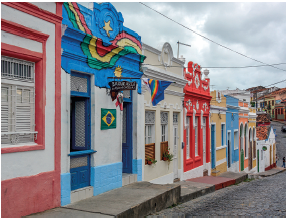 Imagem: Fotografia. À esquerda, calçada no alto descendo com casas de paredes coloridas em branco com partes em vermelho, azul-claro, amarelo, laranja e outras cores. Perto de algumas casas, cabos pretos no alto de energia. No alto, céu nublado com nuvens de cor cinza.  Fim da imagem.