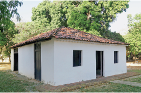 Imagem: Fotografia. Uma casa de paredes brancas, janelas pequenas, portas finas na vertical, com telhado de cor marrom. Ao redor, solo em marrom-claro e grama. Em segundo plano, árvores de folhas verdes. No alto, céu em azul-claro.  Fim da imagem.