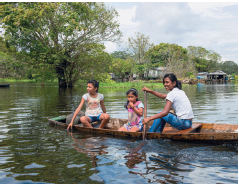 Imagem: Fotografia. Sobre uma canoa de cor marrom, com três meninas. À esquerda, menina de pele morena-claro, cabelos escuros presos atrás, com blusa clara, bermuda azul, com a mão esquerda perto de rio, olhando para frente. Ao centro, menina menor de pele morena, cabelos pretos presos para trás, blusa de mangas curtas em rosa e branco, com a mão direita perto da boca. Na ponta da direita, menina de pele morena, maior que a outras duas, cabelos pretos, com blusa de mangas branca, calça azul, segurando nas mãos, segurando remo marrom. Em segundo plano, árvore de folhas verdes no rio e local com grama verde. Mais ao fundo, casas perto do rio. No alto, céu nublado com nuvens cinza.  Fim da imagem.