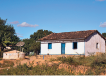 Imagem: Fotografia. Local com solo marrom-claro, vegetação em verde e ao fundo, casa com paredes de cor branca, telhado marrom, com porta azul e janelas pequenas. À esquerda, árvores com folhas verdes.  Fim da imagem.