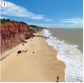 Imagem: Fotografia 1. Vista do alto de local com areia à esquerda, ondas do mar à direita de cor marrom, e à esquerda, areia de cor bege-claro, com morros ao fundo de cor marrom, parte superior com vegetação de cor verde. No alto, céu azul com nuvens em branco.  Fim da imagem.