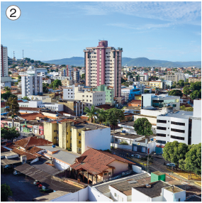 Imagem: Fotografia 2. Visto do alto de local com casas baixas e mais ao fundo, prédios baixos e outros em prédios altos. Em segundo plano, morro alto e no alto, céu em azul-claro, com poucas nuvens brancas esparsas.  Fim da imagem.