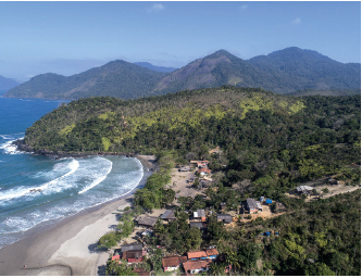 Imagem: Fotografia. Vista geral de local com praia à esquerda, de cor azul com ondas em branco. À frente, local com areia bege, com casas e perto delas, vegetação composta com árvores com folhas em verde-escuro, morros com vegetação e mais ao fundo, outros morros. No alto, céu em azul-claro.  Fim da imagem.