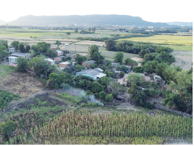 Imagem: Fotografia. Vista geral de local com vegetação de cor verde-claro, ao fundo, local com árvores de folhas em verde-escuro e entre elas, casas de paredes de cor branca com telhados cinzas. Em segundo plano, locais com vegetação verde e morros.  Fim da imagem.