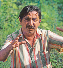 Imagem: Fotografia. Um homem visto da cintura para cima, de pele morena-claro, cabelos e bigode escuros, usando camisa de listras em branco, laranja e verde-claro.  Fim da imagem.