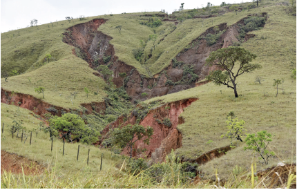 Imagem: Fotografia. Vista geral de local com morros de cor verde e ao centro, fissura ao centro com solo de cor marrom-claro. Sobre o local, algumas árvores de folhas verdes. No alto, céu em azul-claro.  Fim da imagem.