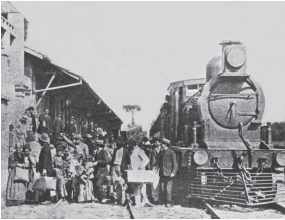 Imagem: Fotografia em preto e branco. À esquerda, estação vista parcialmente com telhado em cima e à frente, centenas de pessoas, homens vestidos de terno e chapéu, mulheres de vestidos longos, carregando malas quadradas antigas. Mais à direita, trem de ferro sobre os trilhos.  Fim da imagem.