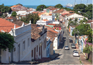 Imagem: Fotografia. Rua em descida ao centro de cor cinza, com carros estacionados à direita e ao centro, poucos carros passando. À esquerda e à direita, casas de paredes coloridas, em bege, azul, laranja, verde e laranja, com muitas janelas. Perto de algumas casas, árvores com folhas verdes. No alto, céu de cor azul-claro e em segundo plano, árvores de folhas em verde-escuro, mar e barcos. Fim da imagem.