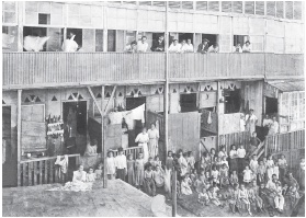 Imagem: Fotografia em preto e branco. moradias em um prédio com inúmeras janelas, em dois andares. No alto, pessoas perto de varanda, atrás de proteção de grade, olhando para frente, homens de terno e alguns com chapéu na cabeça e mulheres de vestes longas claras. Na parte inferior, dezenas de mulheres, homens e crianças nas moradias dispostas lado a lado. Em uma moradia ao centro, roupas penduradas. À frente, na parte inferior, outras pessoas sentadas e em pé perto da grade, com muitas crianças. Alguns adultos têm criança sentada no colo.  Fim da imagem.