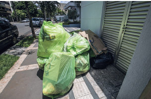 Imagem: Fotografia. Sobre calçada em frente de uma casa de paredes cinzas, três grandes sacolas plásticas de cor verde-claro. Mais ao fundo, outras sacolas menores pretas de plástico, com um papelão de cor marrom em cima. Mais à esquerda, local com vegetação em verde, carro estacionado e ao atrás, árvores de troncos finos, folhas verdes e outras residências.  Fim da imagem.