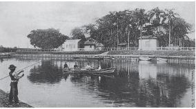 Imagem: Fotografia em preto e branco. À esquerda, um homem em pé de pele escura, camisa de mangas compridas clara, colete e calça escura, com um chapéu sobre a cabeça, segurando no braço esquerdo: vara de pesca. Mais ao fundo, rio grande com uma canoa na horizontal com quatro pessoas sentadas dentro. Ao fundo, local com canoa pequena ancoradas. Em segundo plano, local com vegetação e mais ao fundo, casas antigas e local com muitas árvores com folhas no alto.  Fim da imagem.