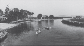 Imagem: Fotografia em preto e branco. Vista do alto de local com rio extenso e duas pessoas sobre ele, dentro de canoa pequena. Mais à esquerda, um barco pequeno visto parcialmente. À esquerda, local com árvores à esquerda e partes com vegetação à esquerda e à direita.  Fim da imagem.