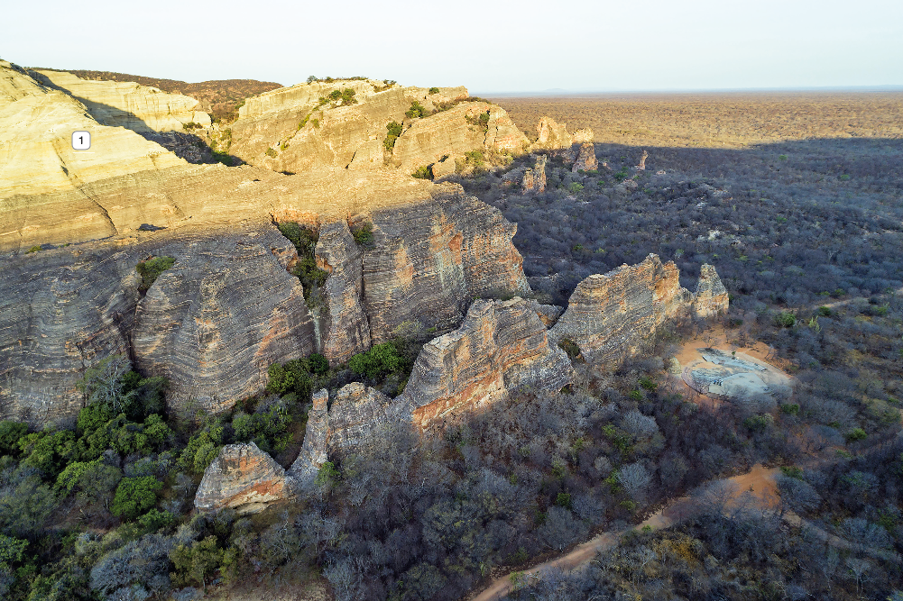 Imagem: Fotografia. Vista do alto de local com rochas grandes de cor marrom-claro e partes em cinza, com partes de tamanho médio e outras grandes com partes com vegetação verde. À direita, local mais baixo, parte com solo bege e vegetação rasteira seca. Em segundo plano, solo com vegetação seca, sem rochas. No alto, céu em azul-claro.  Fim da imagem.